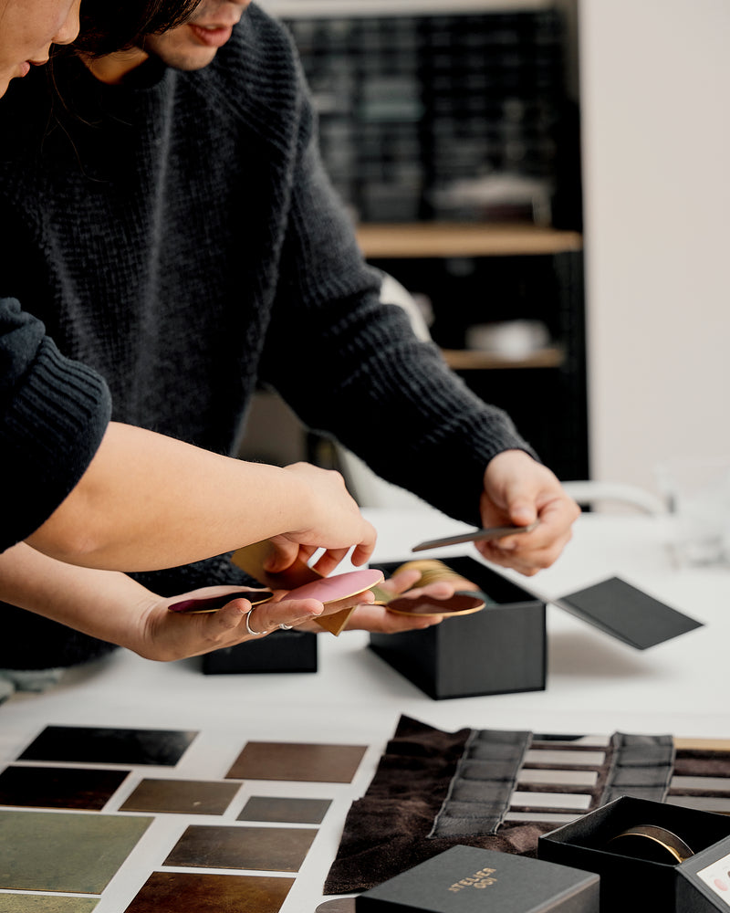 Two people examining small black boxes on a table with color swatches in Atelier001